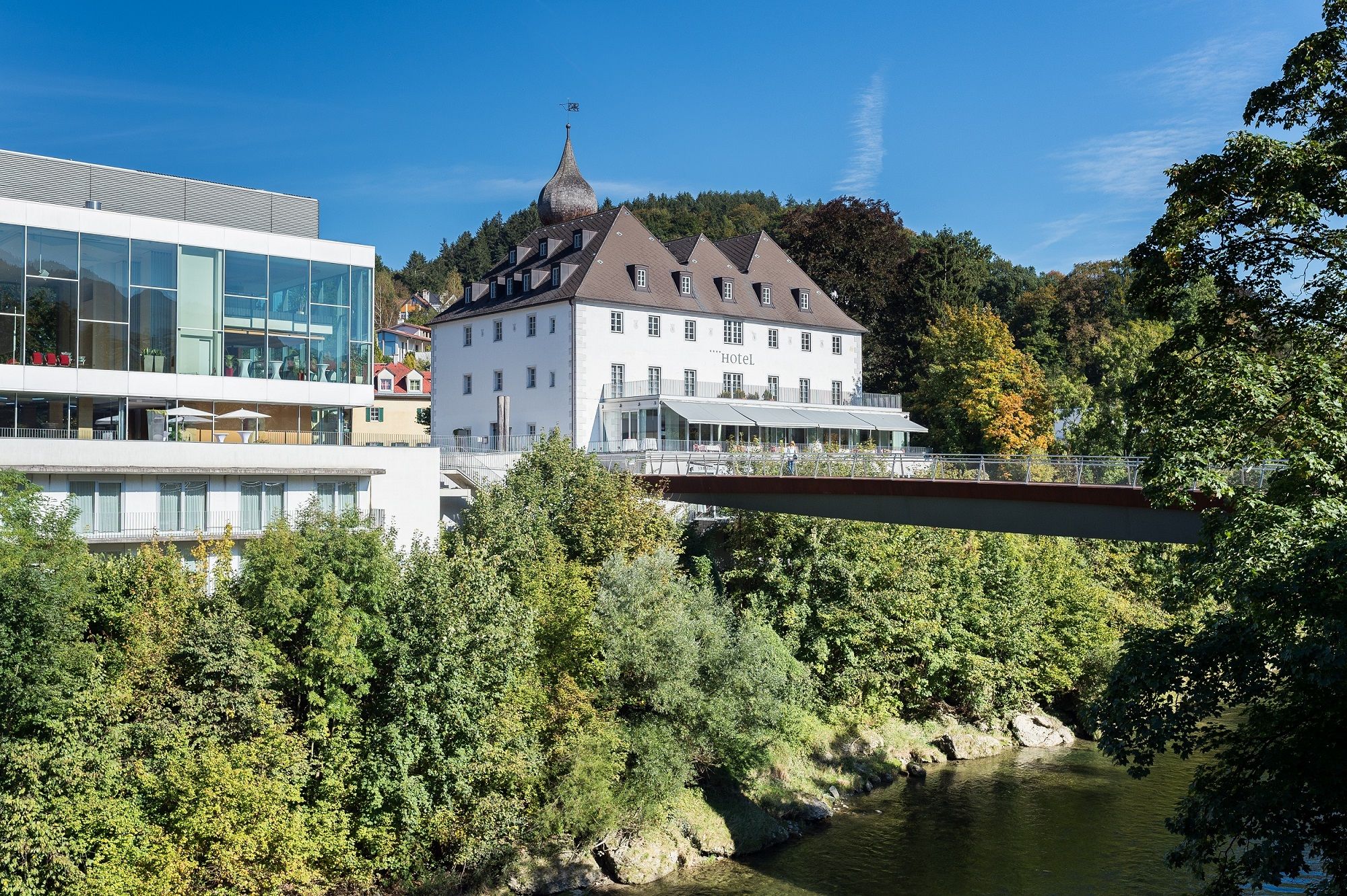 Ein modernes Gebäude neben einem historischen Schloss, umgeben von Bäumen und einem Fluss, unter blauem Himmel.