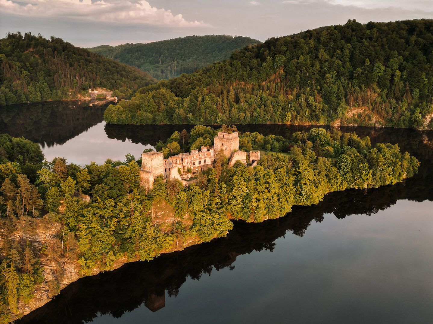 Eine Luftaufnahme der Burgruine Dobra im Waldviertel, umgeben von dicht bewaldeten Hügeln und einem See bei Sonnenuntergang.