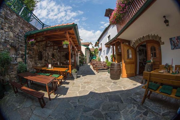 Ein gem&uuml;tlicher Innenhof mit Holzb&auml;nken, Tischen und Blumen. Steinboden und &uuml;berdachte Sitzbereiche. Sonniges Wetter mit blauem Himmel.