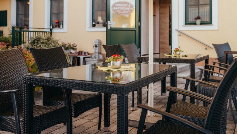 Gem&uuml;tlicher Au&szlig;enbereich eines Gasthauses mit Rattanm&ouml;beln und Blumen.