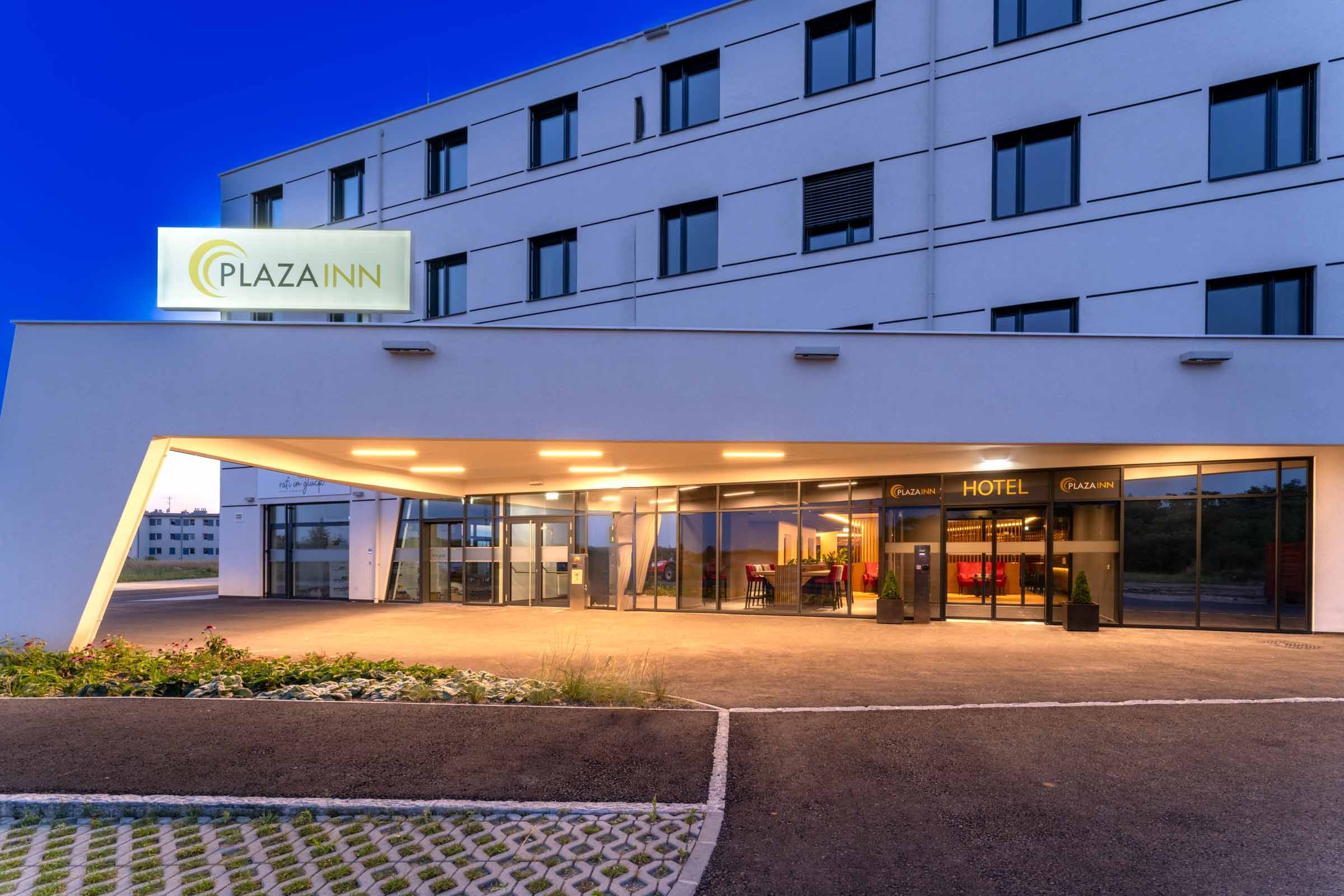 Eingang des Plaza Inn Hotels bei Nacht mit beleuchtetem Schild und moderner Architektur.