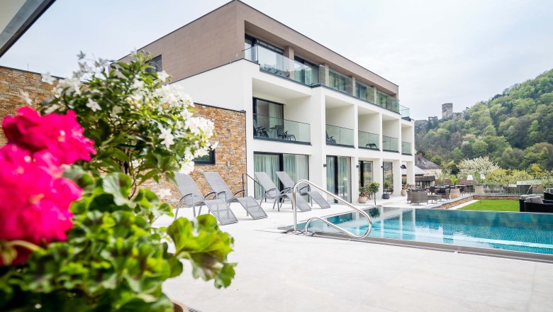Modernes Gebäude mit Pool und Liegestühlen, umgeben von grüner Landschaft und Burg im Hintergrund.