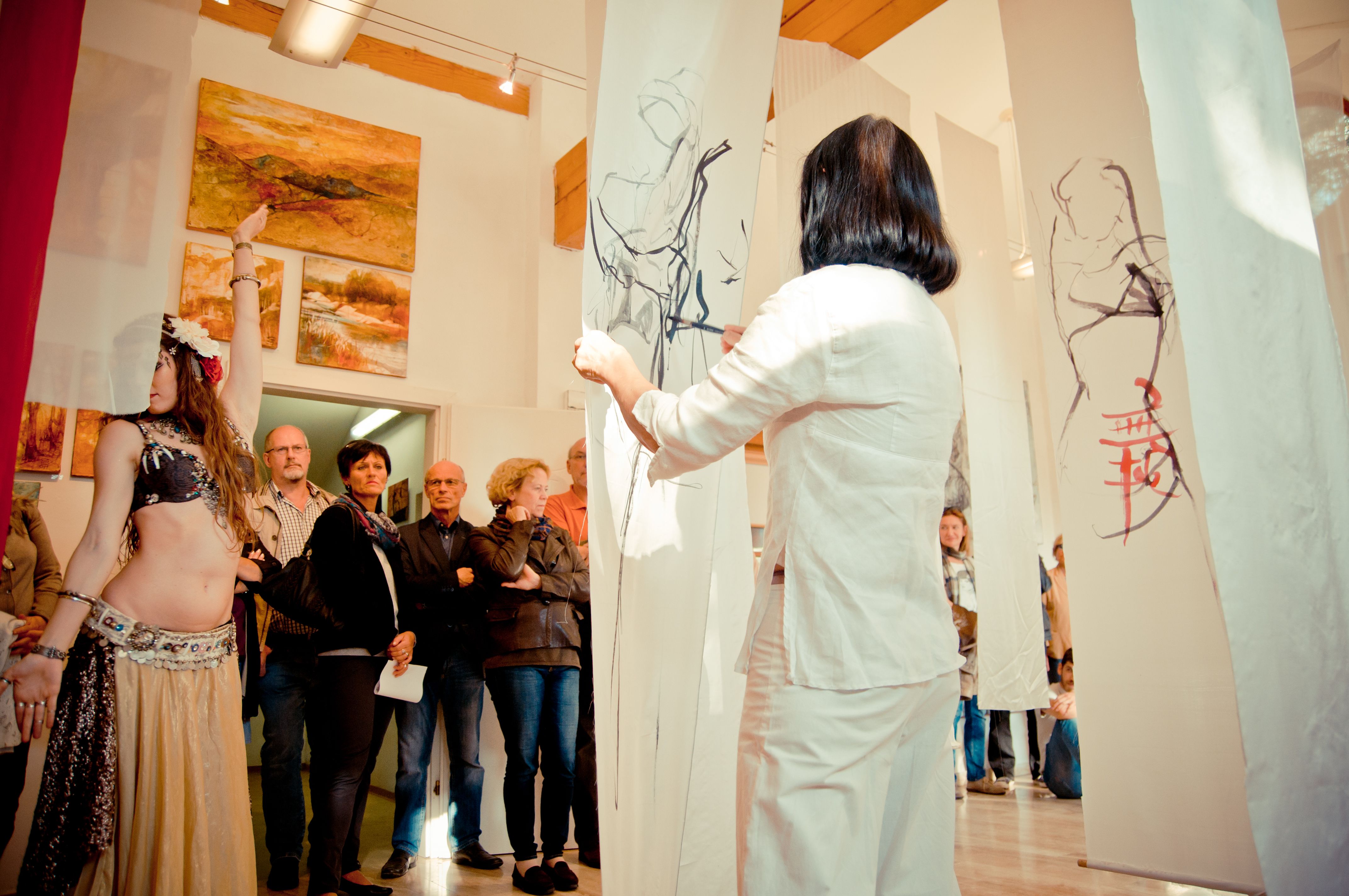 Eine Frau in weißer Kleidung betrachtet Zeichnungen auf hängenden Stoffbahnen in einer Galerie mit Zuschauern und Gemälden an der Wand.