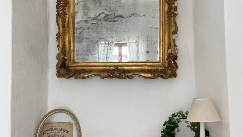 Ein antiker Spiegel mit goldener Verzierung über einer Fensterbank mit Deko.