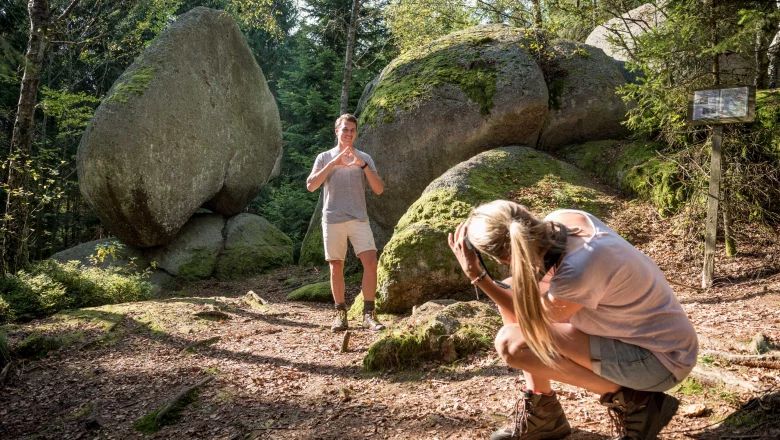 Ein Mann posiert vor einem großen herzförmigen Felsen im Wald, während eine Frau ein Foto von ihm macht.
