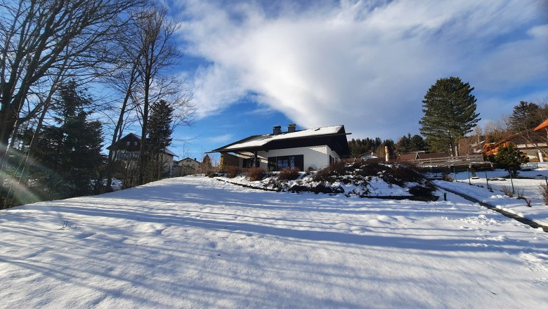Winterlandschaft mit einem Haus im Schnee, umgeben von B&auml;umen und blauem Himmel.