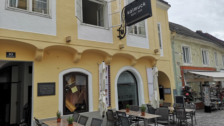 Außenansicht eines gelben Gebäudes mit dem Schild 'Kalmuck Wein Bar'. Vor dem Gebäude stehen Tische und Stühle.