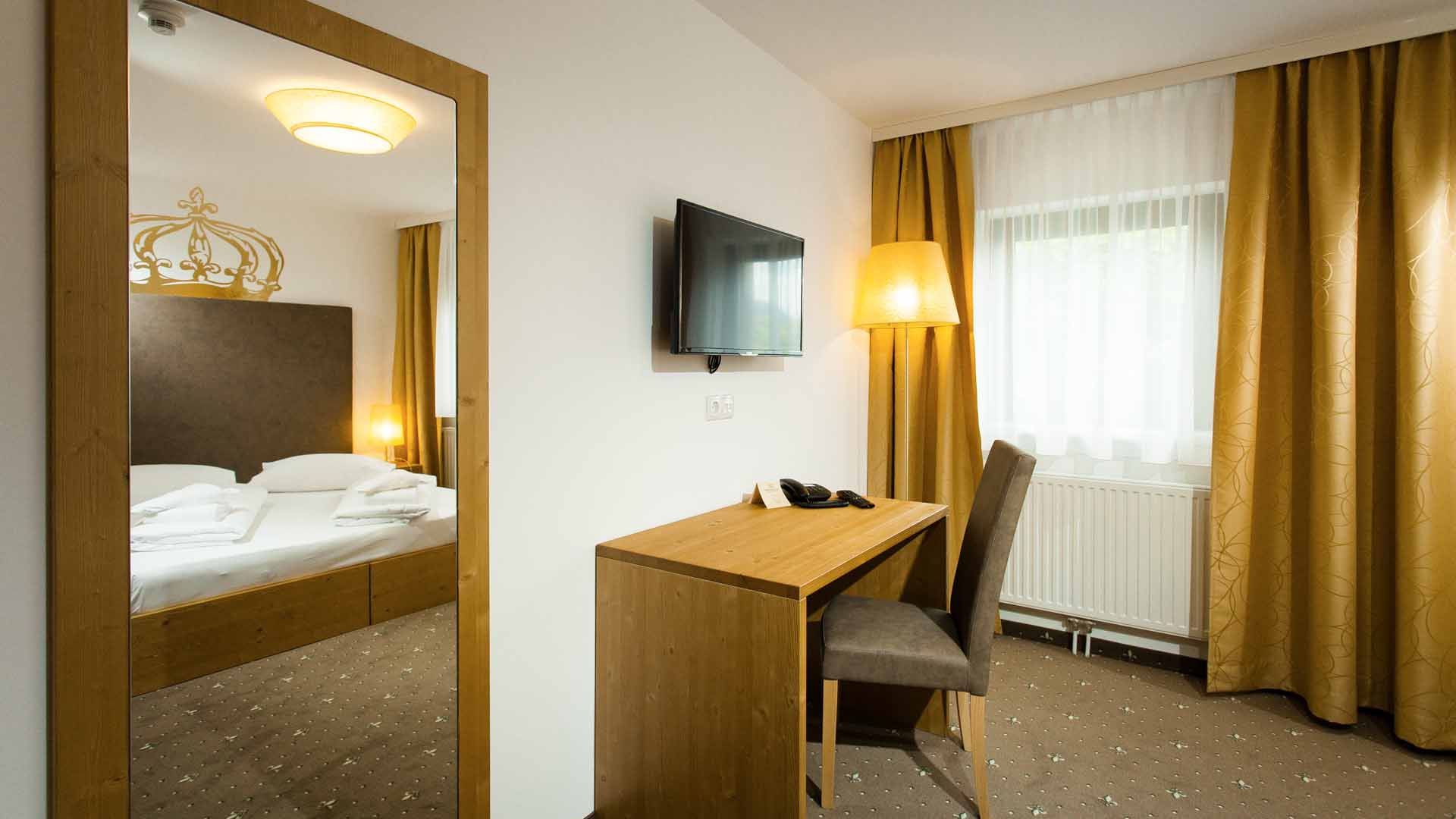 Ein Hotelzimmer mit einem Doppelbett, einem Schreibtisch mit Stuhl, einem Fernseher an der Wand und gelben Vorhängen.