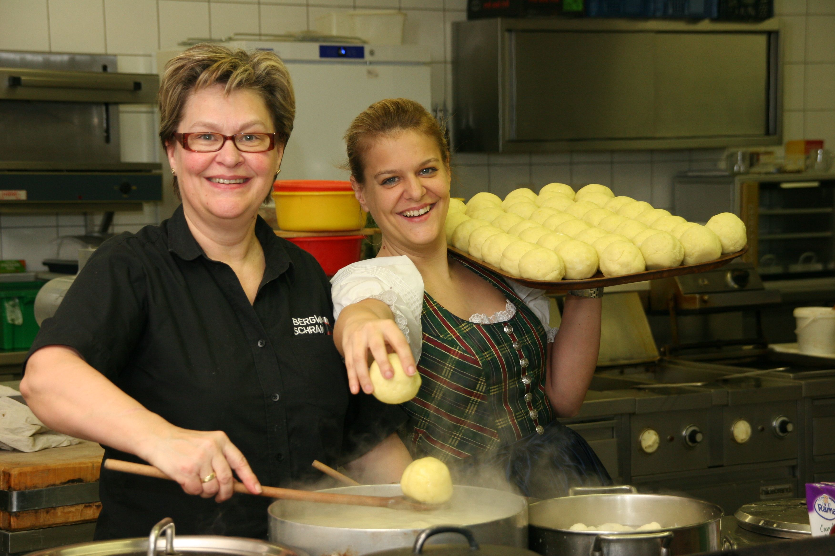 Zwei Frauen in einer Küche, eine hält einen Tablett mit Knödeln, die andere kocht Knödel in einem Topf.