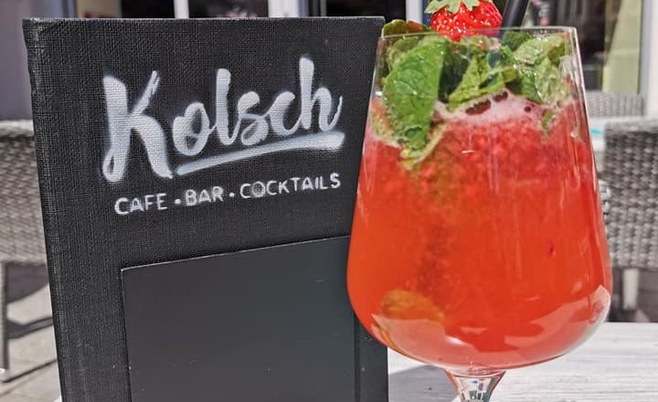 Ein roter Cocktail mit Minze und Erdbeere vor einem Café mit Schild 'Kölsch Café Bar Cocktails'.