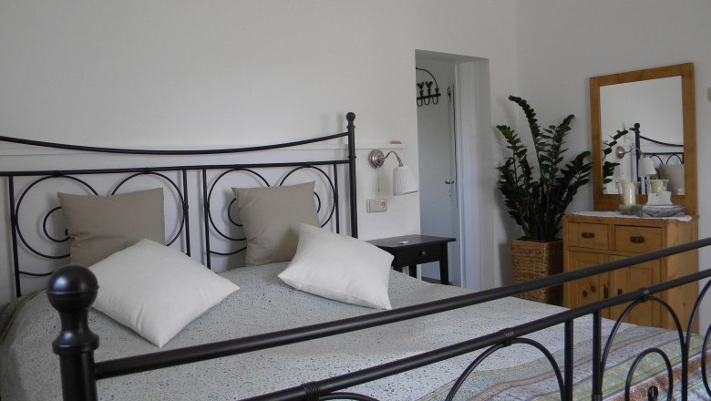 Schlafzimmer mit Metallbett, Kommode und Pflanze.