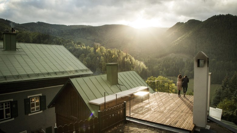 Zwei Personen stehen auf einer Terrasse mit Blick auf bewaldete Berge bei Sonnenuntergang.
