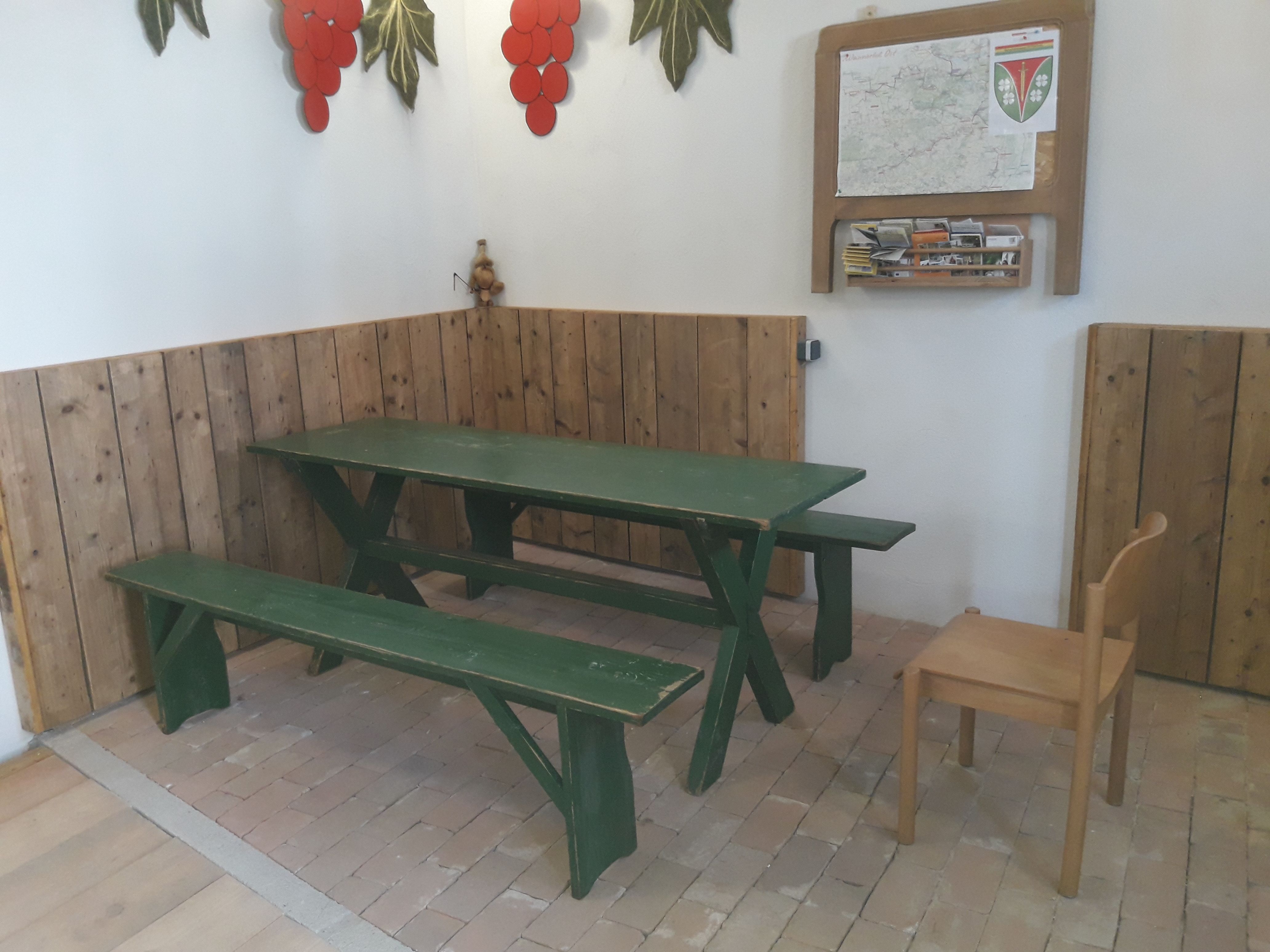 Rustikaler Raum mit grünem Holztisch und Bänken, dekoriert mit Weintraubenmotiven an der Wand.