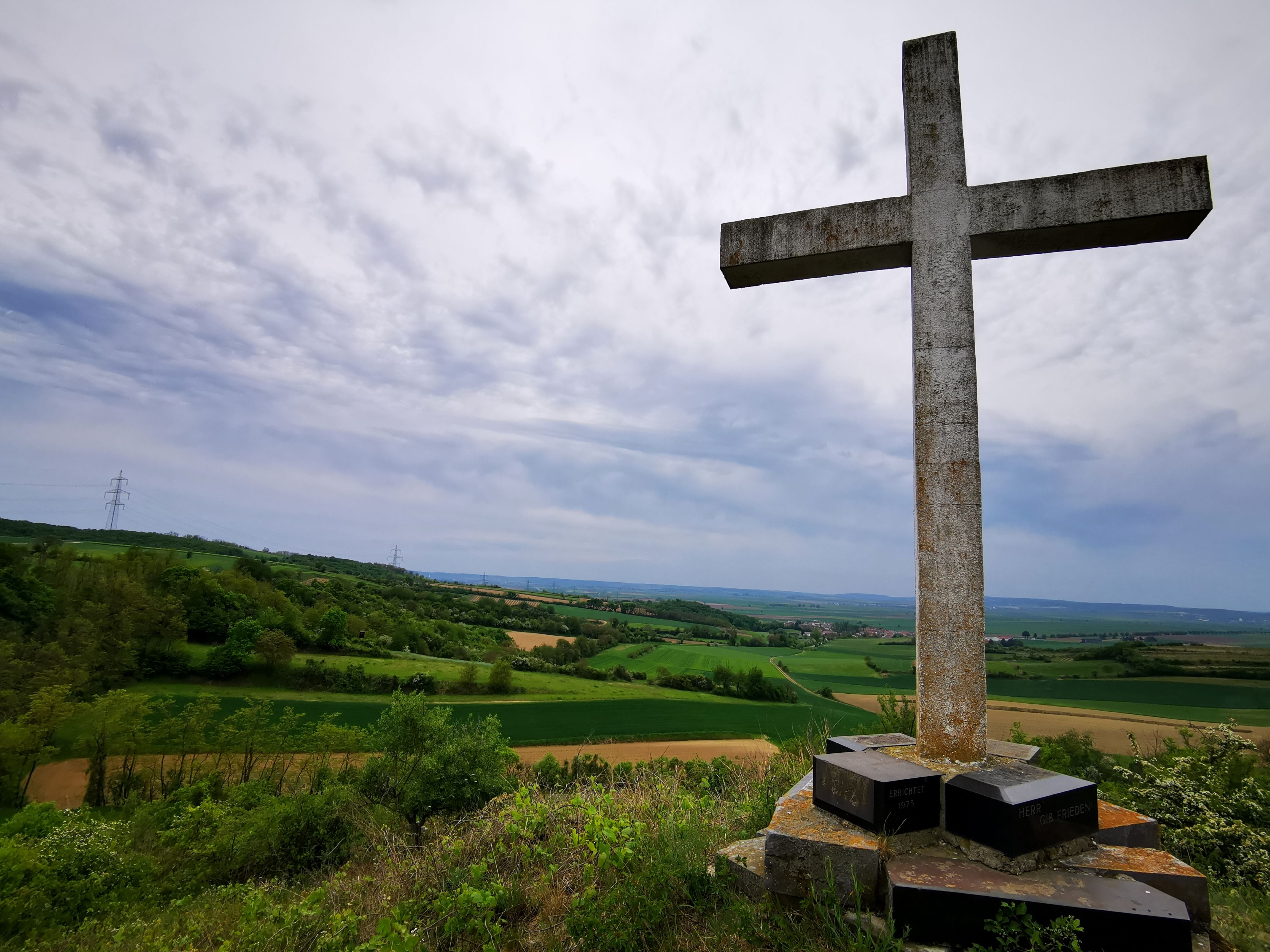 Ein großes Steinkreuz auf einem Hügel mit Blick auf eine grüne Landschaft und bewölkten Himmel.