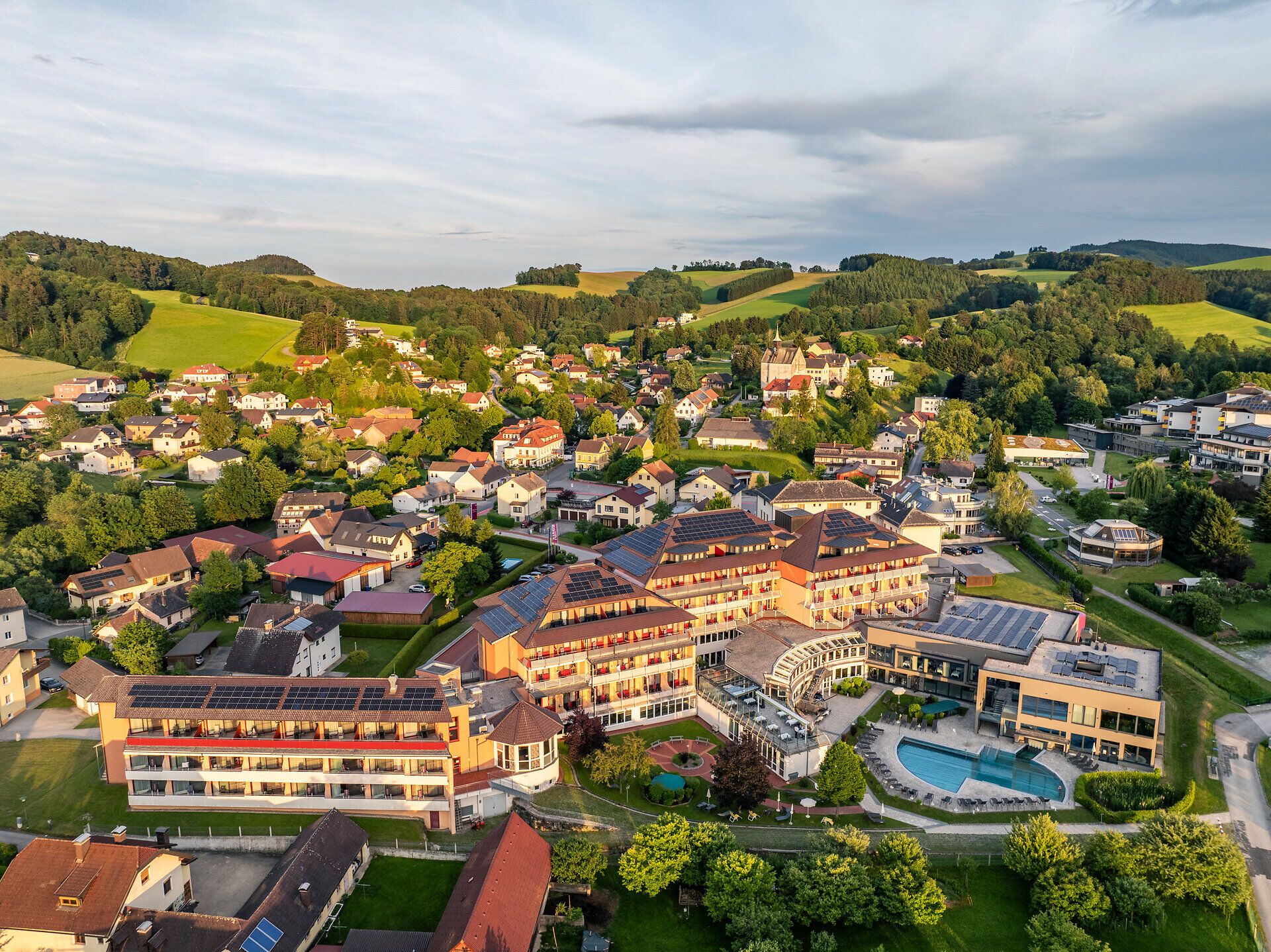 Luftansicht auf einen Ort mit großem Hotel im Bildfokus inmitten von einer Hügellandschaft aus Wiesen, Feldern und Wäldern.