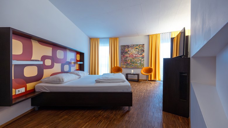 Modernes Hotelzimmer mit Doppelbett, buntem Wandmuster, zwei orangefarbenen Stühlen und gelben Vorhängen.