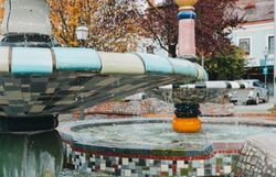 Der Herbst verleiht dem Hundertwasserbrunnen eine besondere Magie, während das Wasser sanft über die bunten Fliesen plätschert. Umgeben von leuchtenden Bäumen und einer einladenden Atmosphäre, lädt dieser Ort zum Verweilen und Träumen ein.