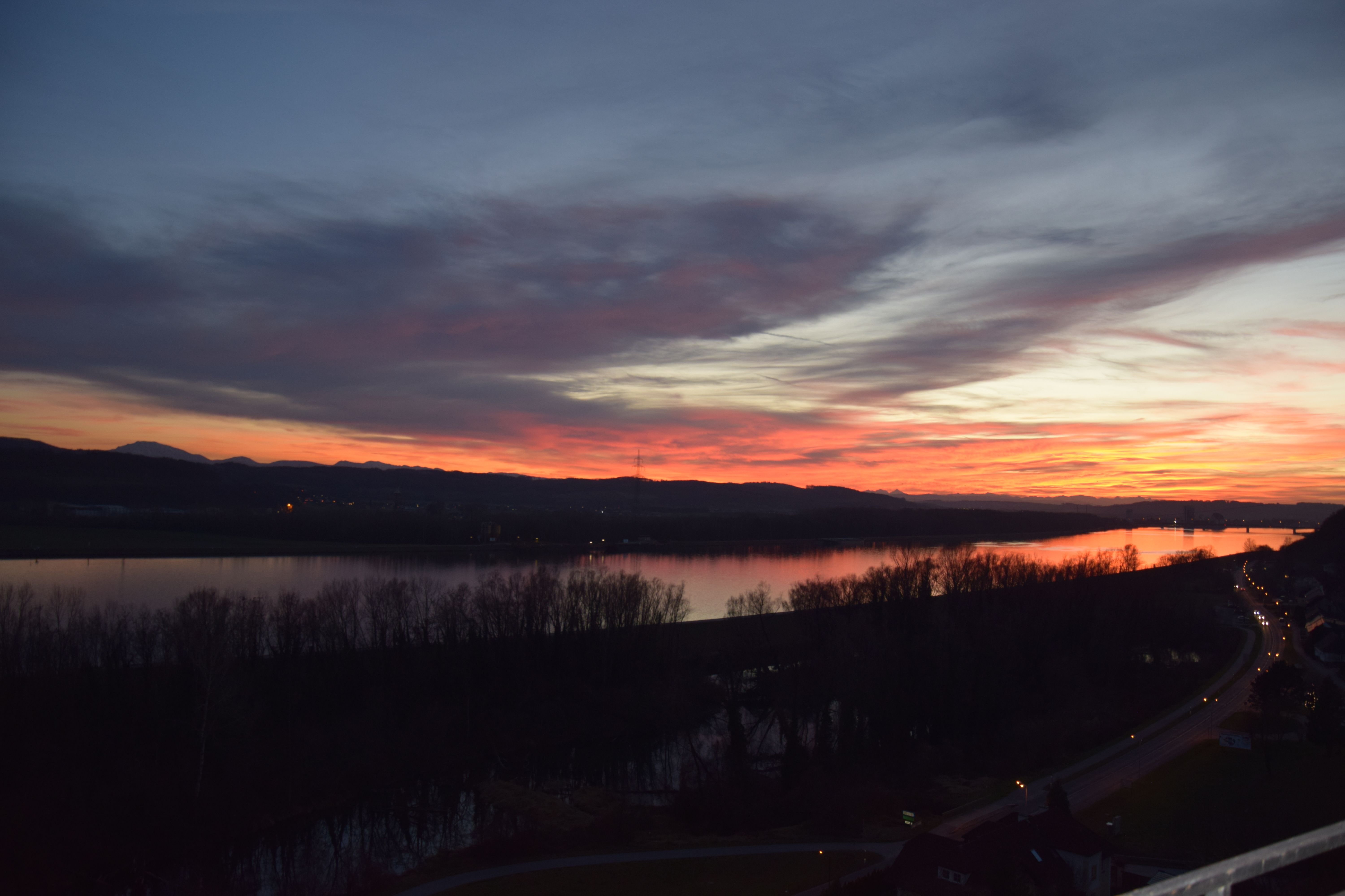 Sonnenuntergang über einem Fluss mit Bäumen und einer Straße im Vordergrund.