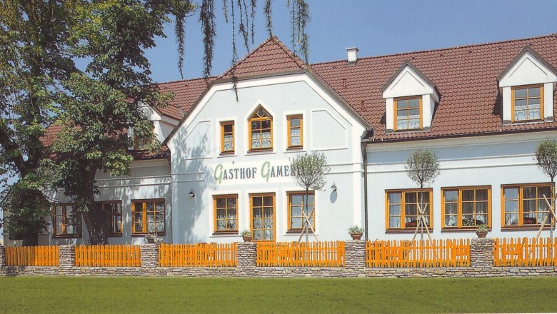 Ein traditionelles Gasthaus mit wei&szlig;er Fassade und roten Dachziegeln, umgeben von B&auml;umen und einem Holzzaun.