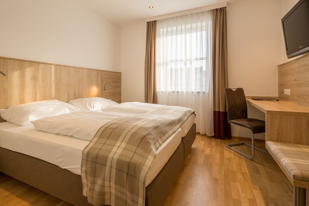 Hotelzimmer mit Doppelbett, Schreibtisch und Fenster mit Vorhängen.