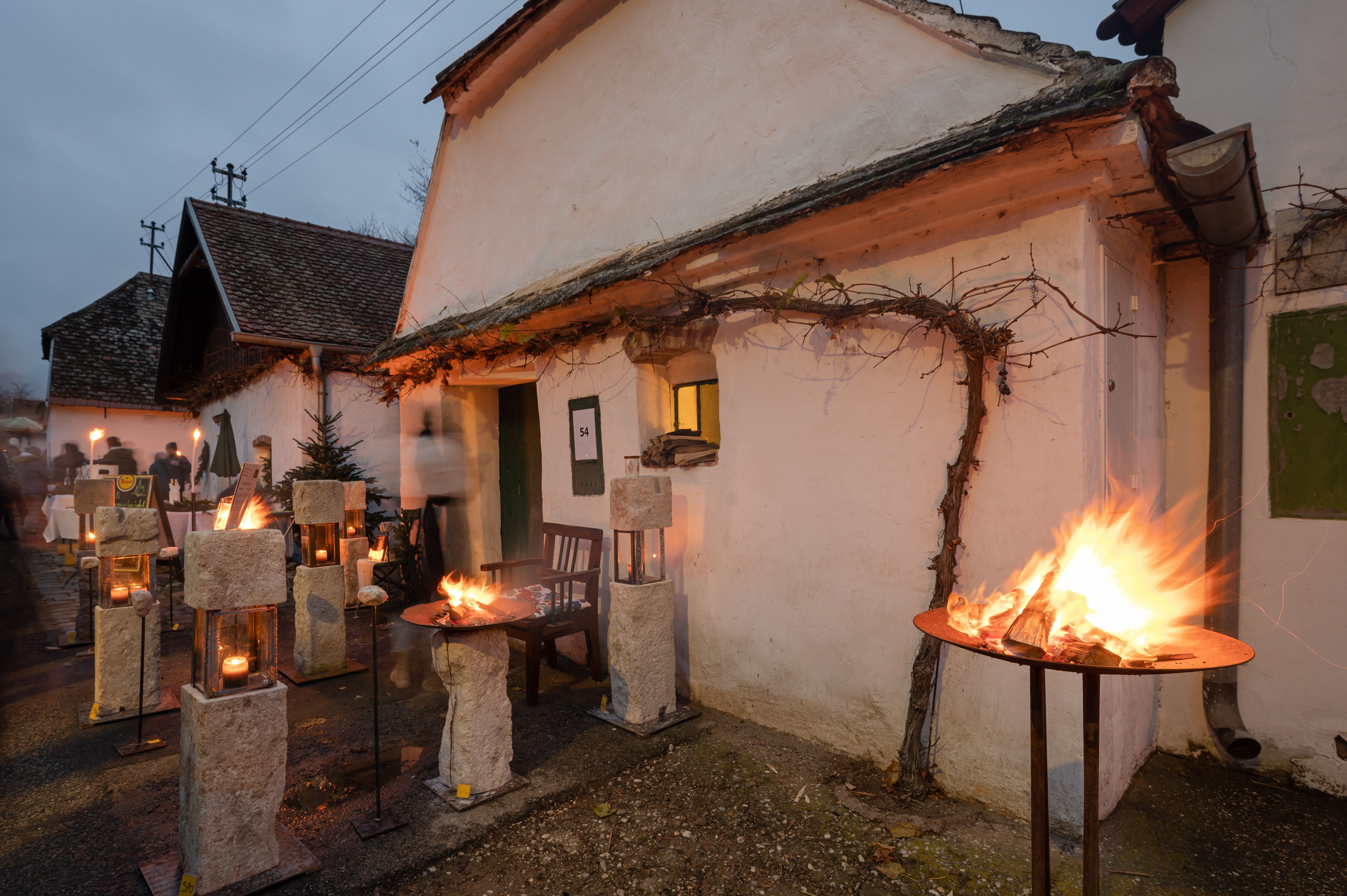 Advent in Hadres: beleuchteter Hof mit Feuerkörben, Laternen und rustikalen Häusern bei Dämmerung.