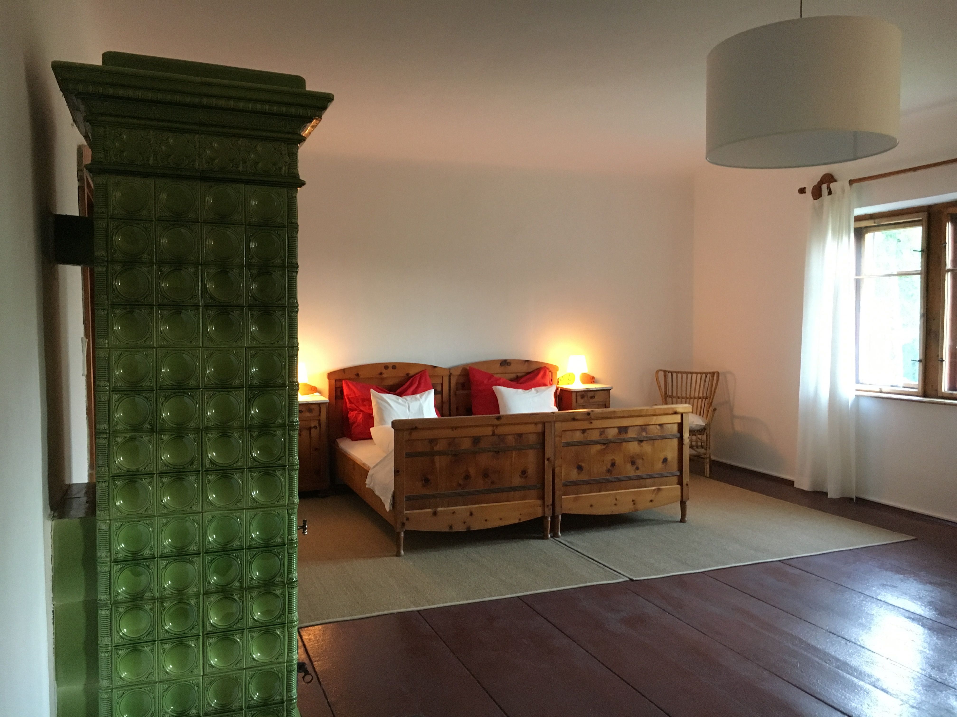 Ein gemütliches Doppelzimmer mit Holzmöbeln, einem grünen Kachelofen und einem großen Fenster mit weißen Vorhängen.