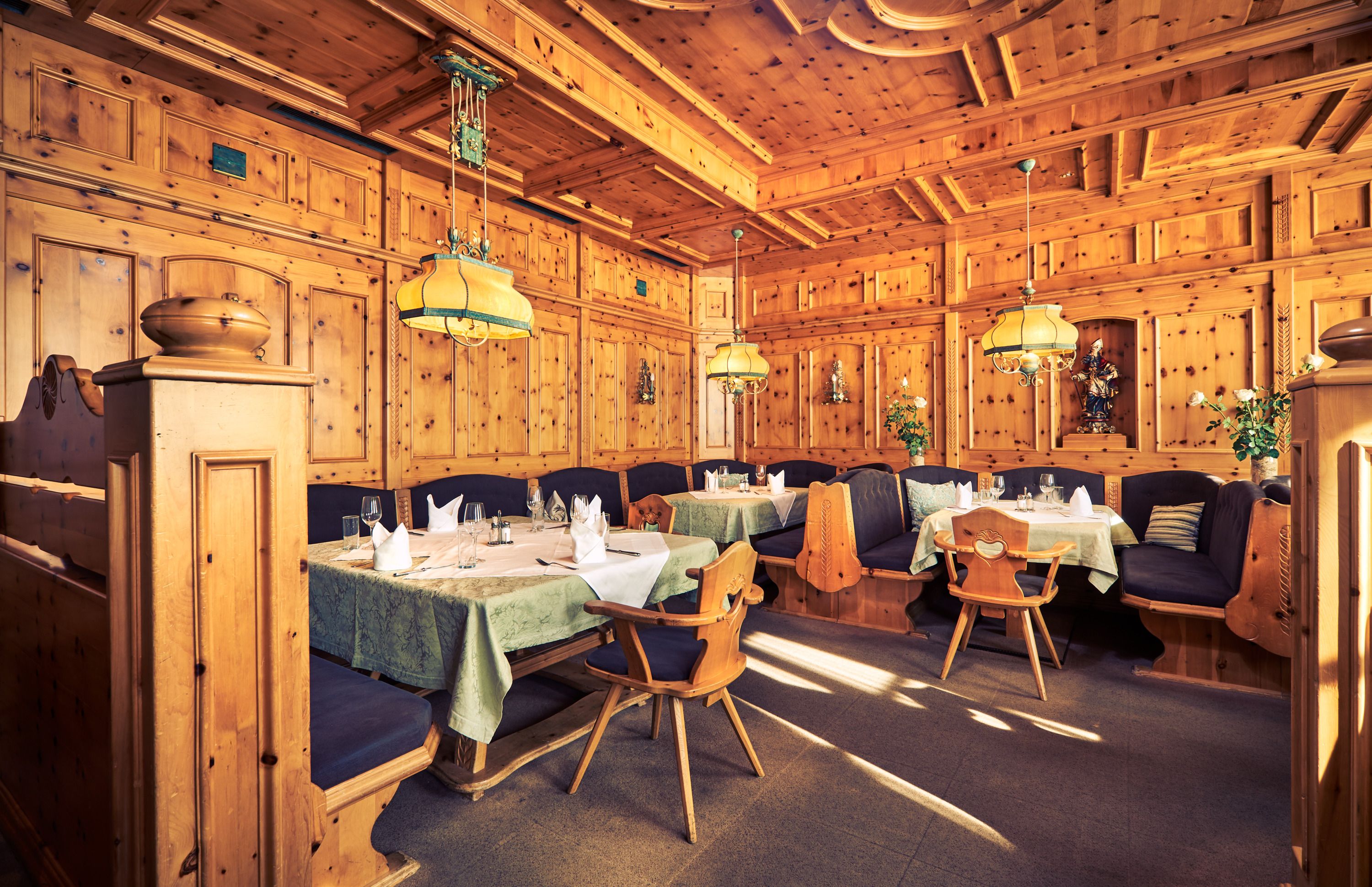 Gemütliche Zirbenstube mit Holzwänden, Tischen und Stühlen, dekoriert mit Lampen und Pflanzen.