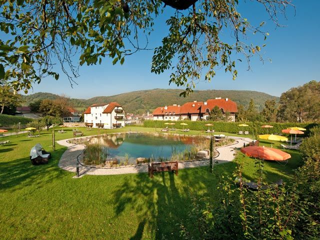 Gartenanlage mit Teich, Liegestühlen und Sonnenschirmen, umgeben von Gebäuden und Hügeln.