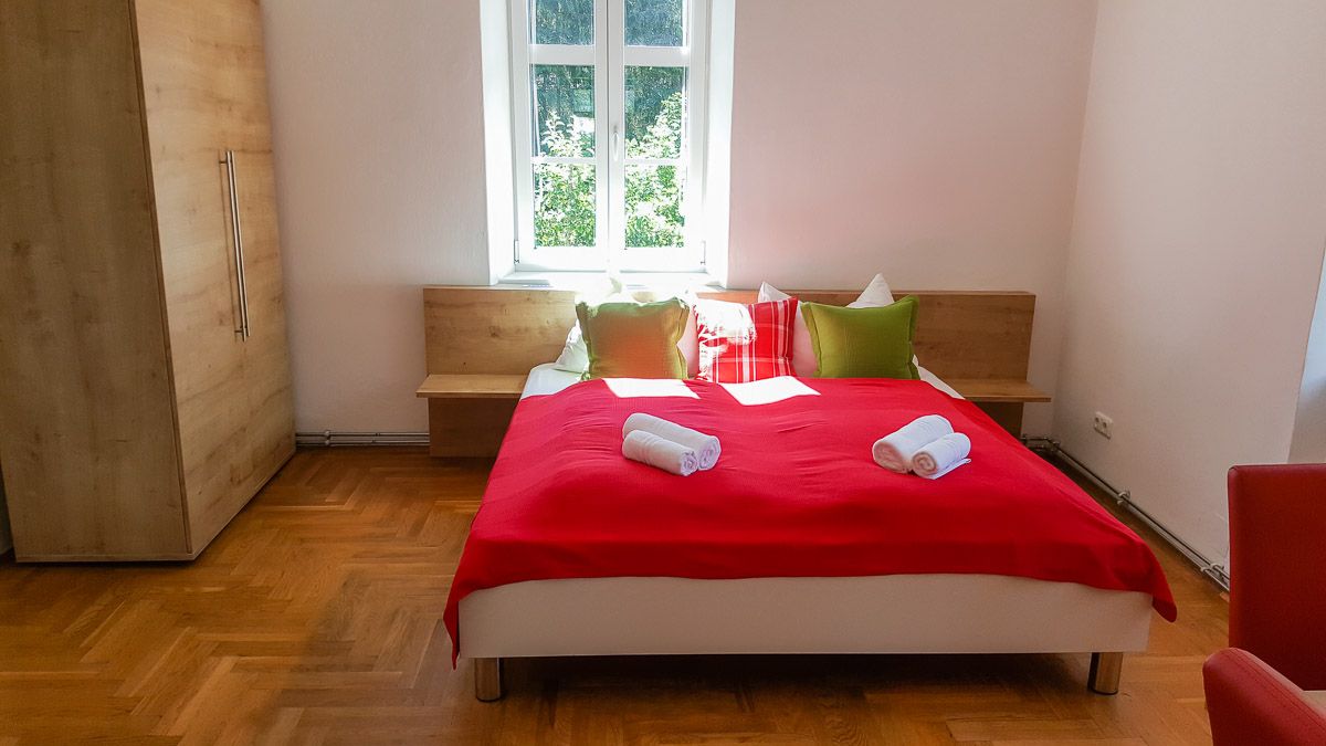 Schlafzimmer mit rotem Bettüberwurf, Kissen und Handtüchern auf dem Bett, Holzschrank und Fenster im Hintergrund.