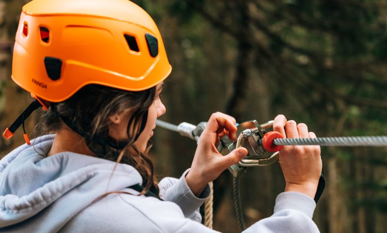 Person mit orangefarbenem Helm befestigt Sicherheitsausrüstung an einem Seil im Waldseilgarten.