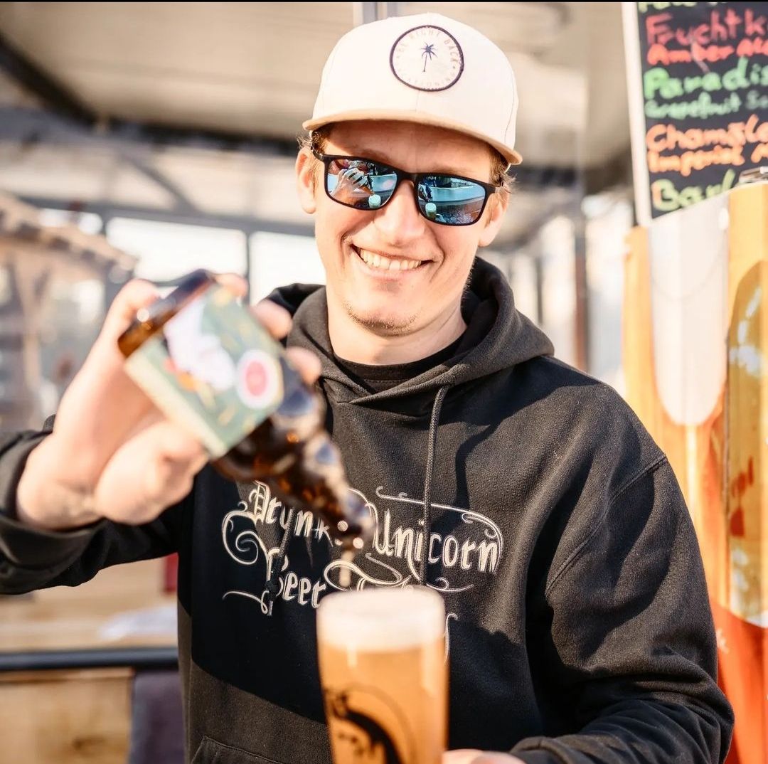 Mann mit Sonnenbrille und Mütze schenkt Bier aus einer Flasche in ein Glas ein.