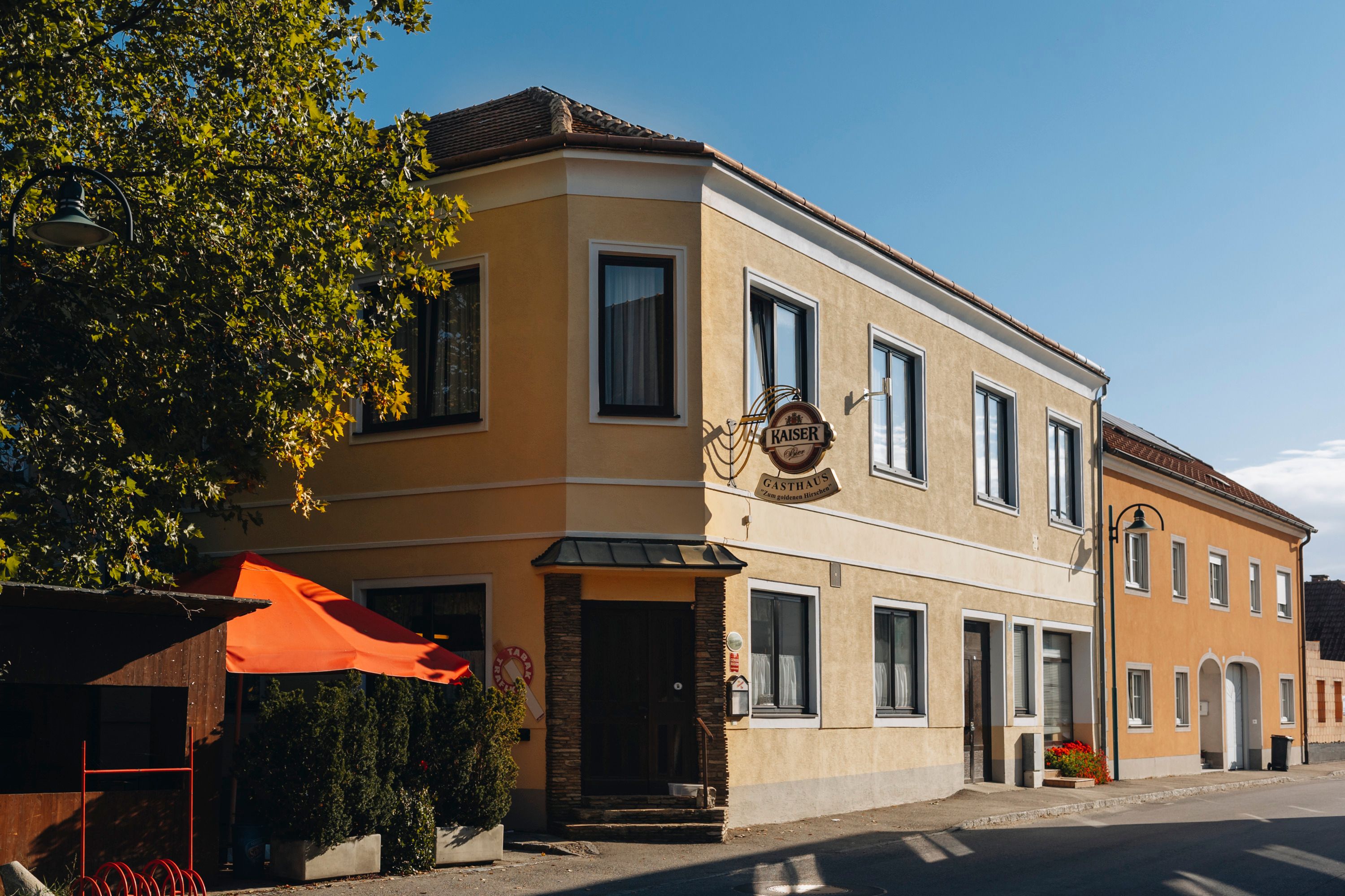 Gelbes Gasthaus mit Schild und rotem Sonnenschirm in sonniger Straße.