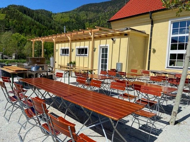 Biergarten mit Holztischen und Stühlen vor einem gelben Gebäude.