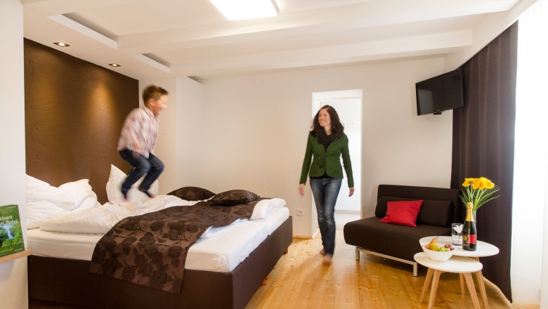 Ein Kind springt auf einem Bett in einem modernen Hotelzimmer, w&auml;hrend eine Frau l&auml;chelnd zusieht. Ein Tisch mit Blumen und Wein steht daneben.