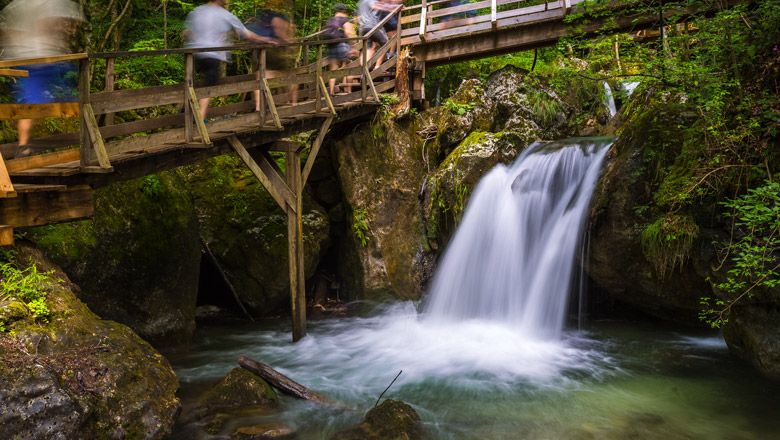 Holzbrücke über Wasserfall in bewaldeter Umgebung.