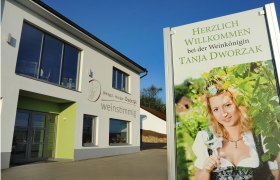 Ein modernes Geb&auml;ude mit einem Schild, das eine Frau in Tracht mit einem Weinglas zeigt. Text: Herzlich willkommen bei der Weink&ouml;nigin Tanja Dworzak.