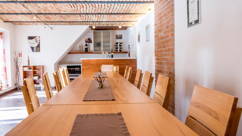 Verkostungsraum mit langem Holztisch und St&uuml;hlen, Ziegeldecke und Weinregal.