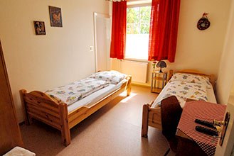 Ein einfach eingerichtetes Zimmer mit zwei Einzelbetten, einem Fenster mit roten Vorh&auml;ngen und einem kleinen Tisch mit Stuhl.