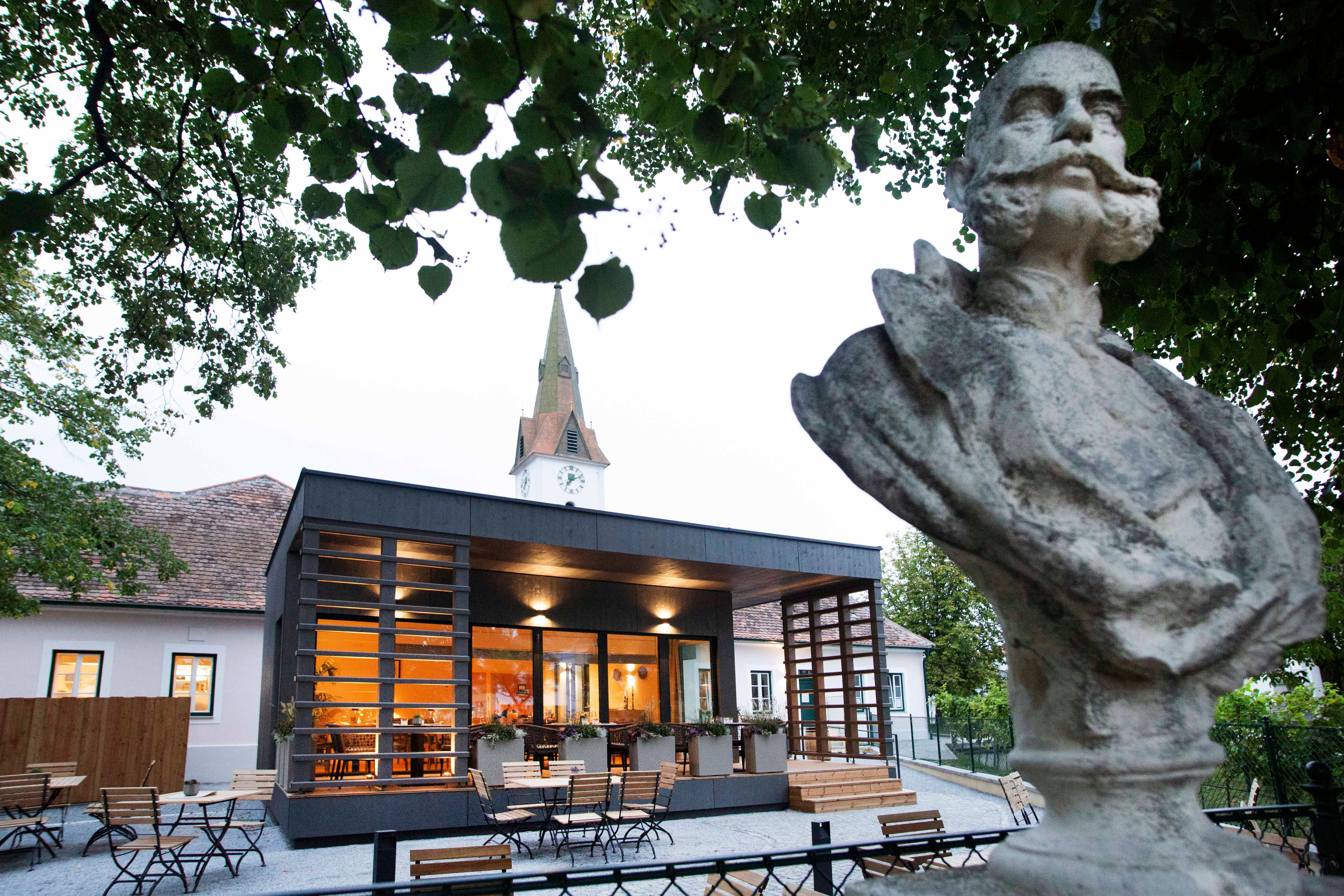 Büste vor modernem Gebäude mit Kirchturm im Hintergrund, umgeben von Bäumen.