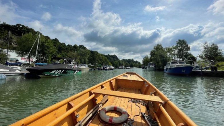 Blick von einem Holzboot auf einen Hafen mit mehreren Booten und Bäumen am Ufer unter einem bewölkten Himmel.