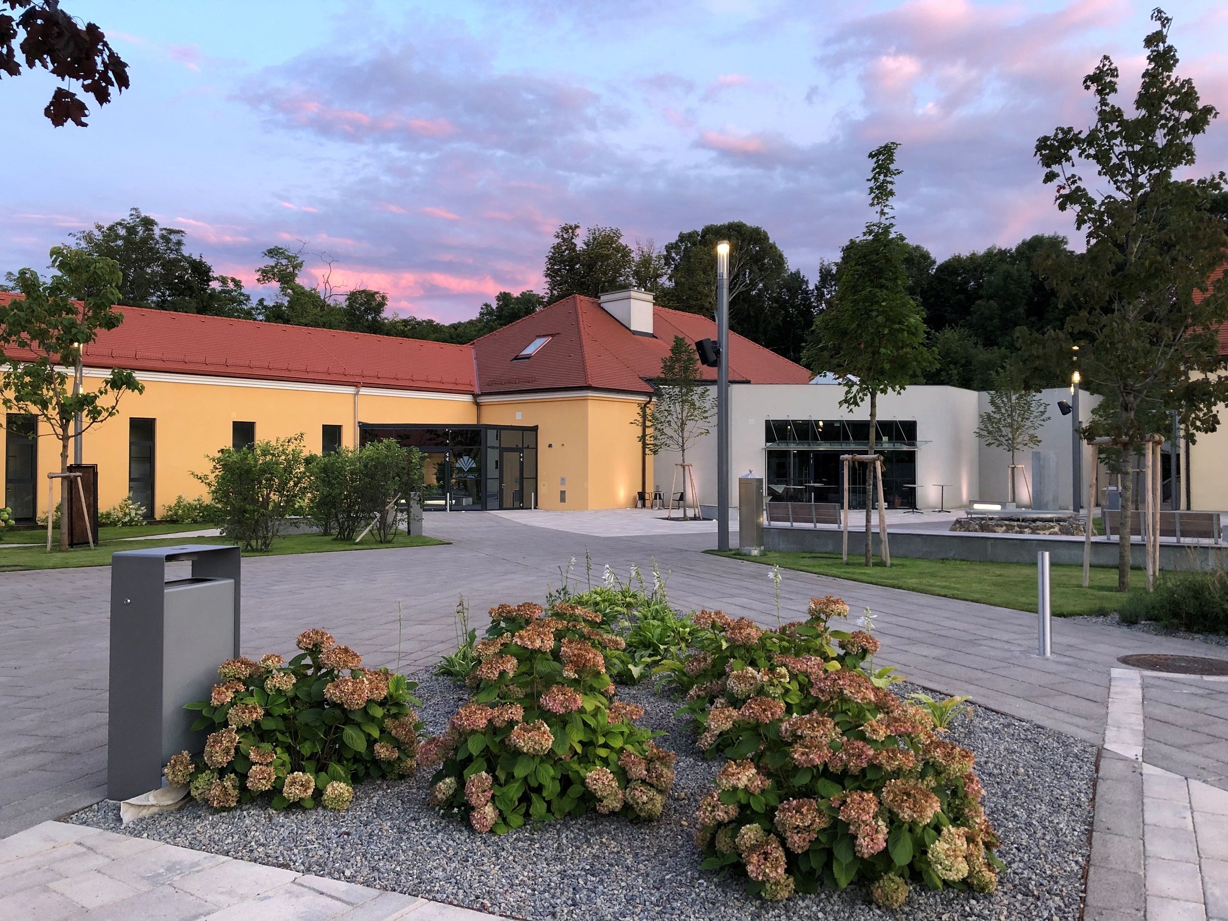 Modernes Gebäude mit gelber Fassade und rotem Dach, umgeben von Pflanzen und Bäumen, bei Sonnenuntergang.