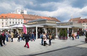 Menschenmenge auf einem belebten Marktplatz mit Ständen und Gebäuden im Hintergrund.