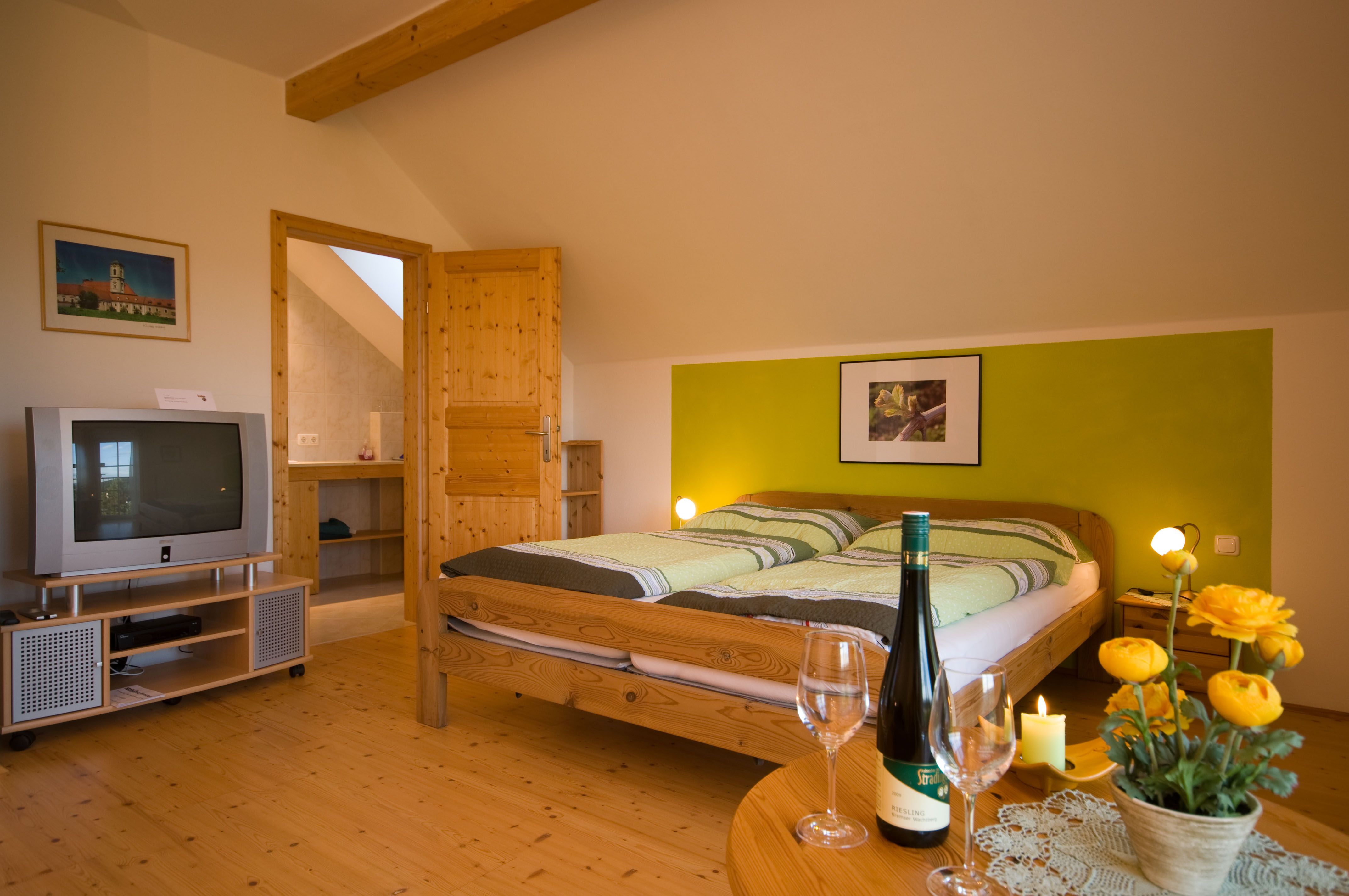 Gemütliches Zimmer mit Doppelbett, TV, Weinflasche und Blumen auf Tisch.