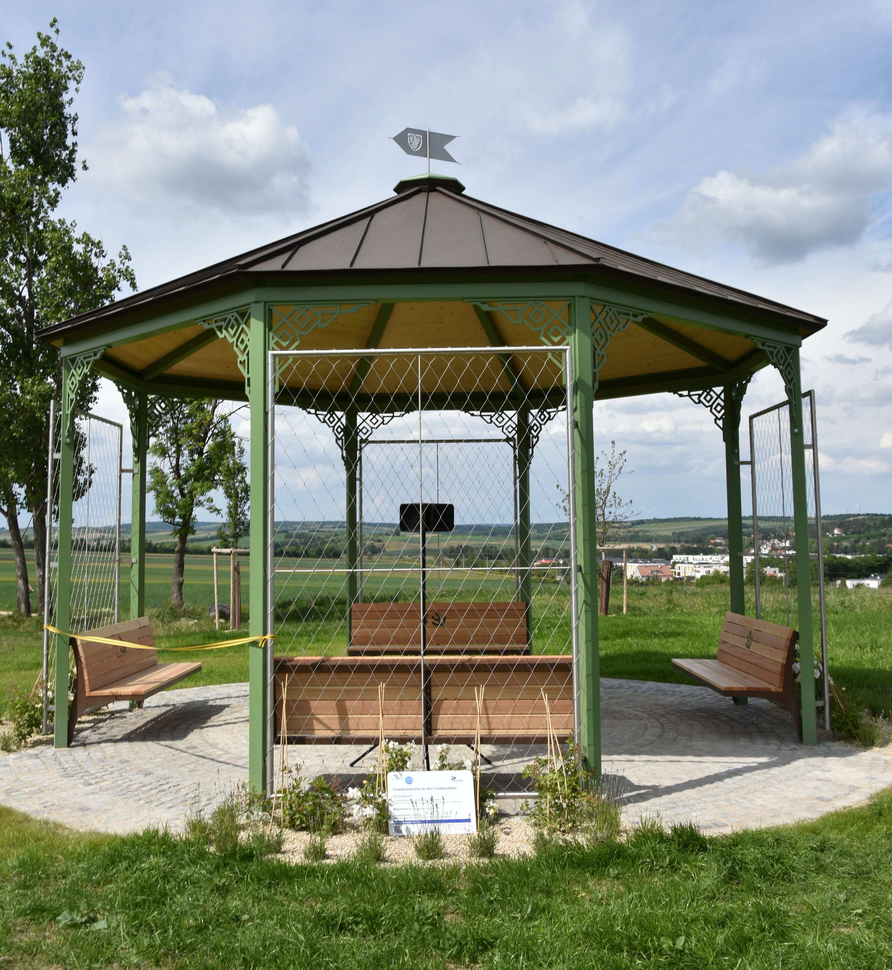 Ein Pavillon mit grünem Metallrahmen und Holzdach steht auf einem gepflasterten Platz in einer ländlichen Umgebung.