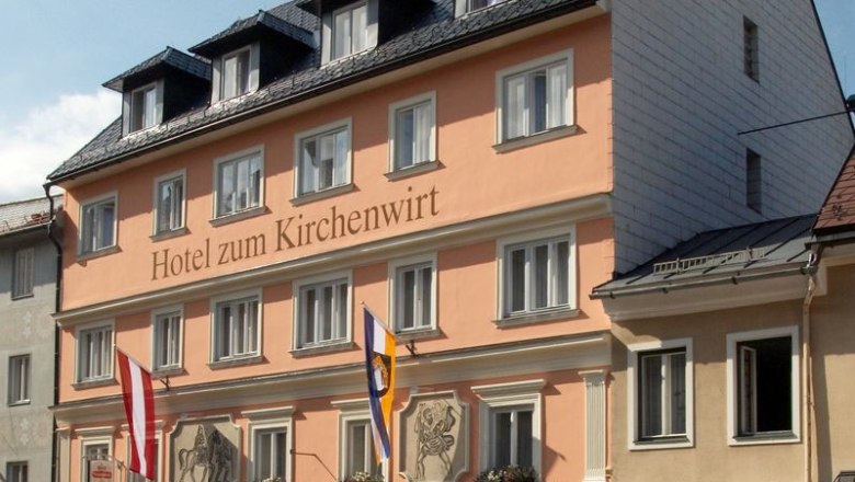 Hotel zum Kirchenwirt, © zVg Hotel zum Kirchenwirt Fassade des Hotels zum Kirchenwirt mit Fahnen und blauer Himmel.