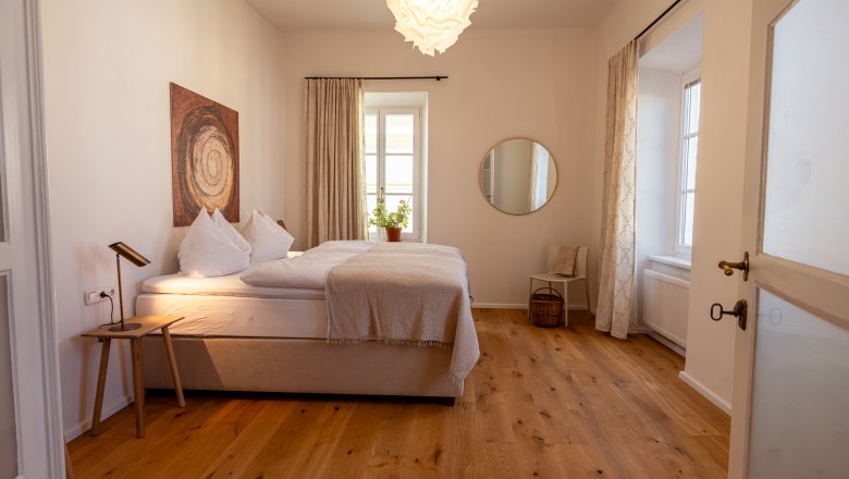 Gem&uuml;tliches Schlafzimmer mit Doppelbett, Holzfu&szlig;boden, Wandkunst, rundem Spiegel und Pflanzen auf dem Fensterbrett.