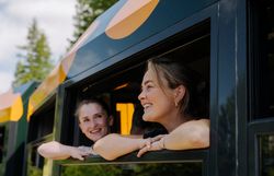 Zwei fröhliche Frauen schauen aus dem Fenster des Salamander-Zuges der Schneebergbahn, während die Landschaft aus Bäumen im Hintergrund vorbeizieht, was Freude und Neugier auf das bevorstehende Reiseerlebnis widerspiegelt.