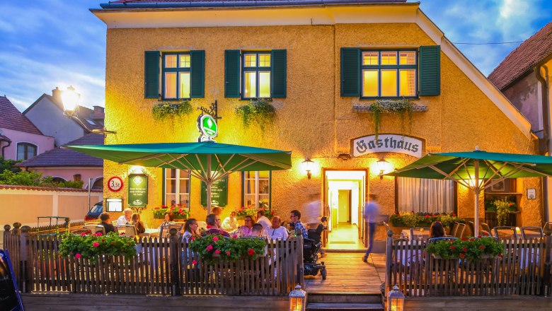 Au&szlig;enansicht eines Gasthauses mit Terrasse und G&auml;sten bei Abendd&auml;mmerung.