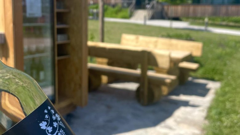 Eine Flasche mit der Aufschrift 'Spritzer rocks' vor einem Rastplatz mit Holzb&auml;nken und einem K&uuml;hlschrank im Freien.