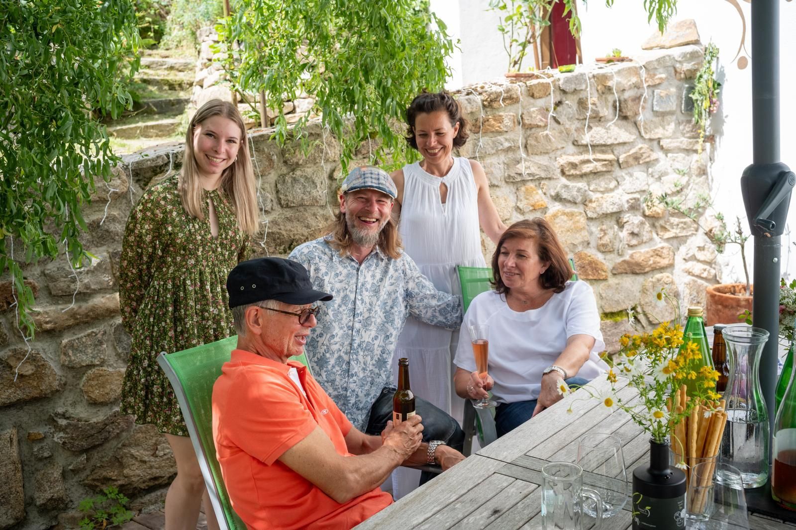 Gruppe von fünf Personen sitzt und steht an einem Tisch im Freien, umgeben von Pflanzen und einer Steinmauer.